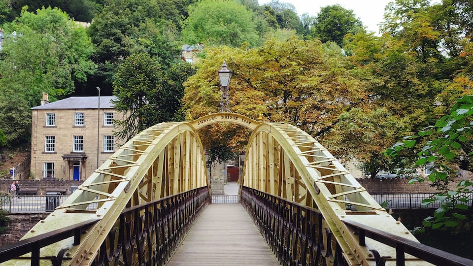 A pedestrian bridge with a sign reading 'Jubilee Bridge 1894' amidst lush greenery and a building in the background.