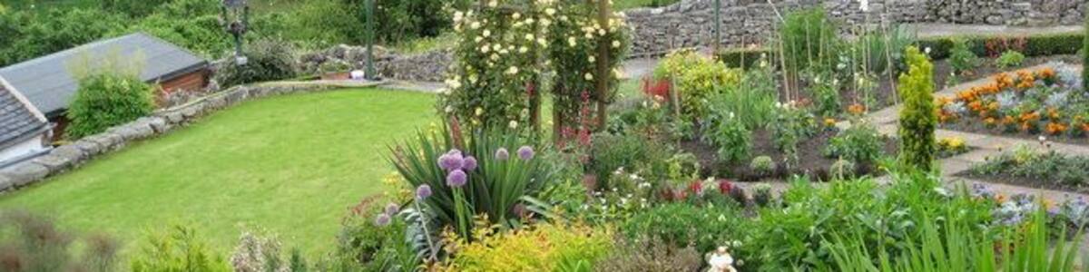 Ein üppiger Garten mit verschiedenen Blumen und Pflanzen, eine Steinmauer und im Hintergrund sanfte Hügel mit traditionellen Steinhäusern.