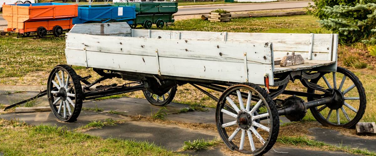 A white wooden wagon with large spoked wheels, parked on a paved surface with grass and a fence in the background.