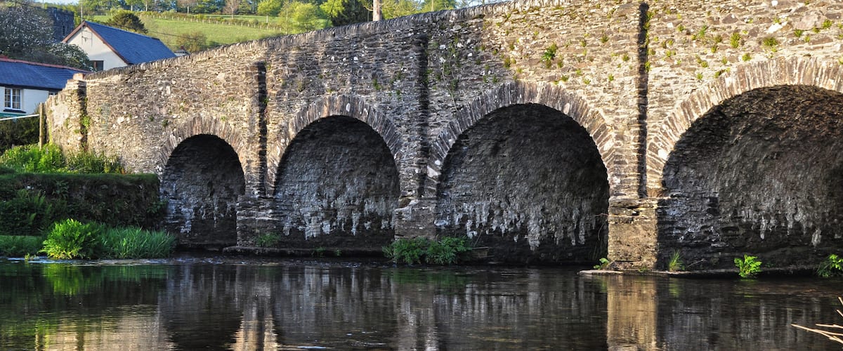 A stone bridge with three arches over a calm river, surrounded by greenery and a house in the background.