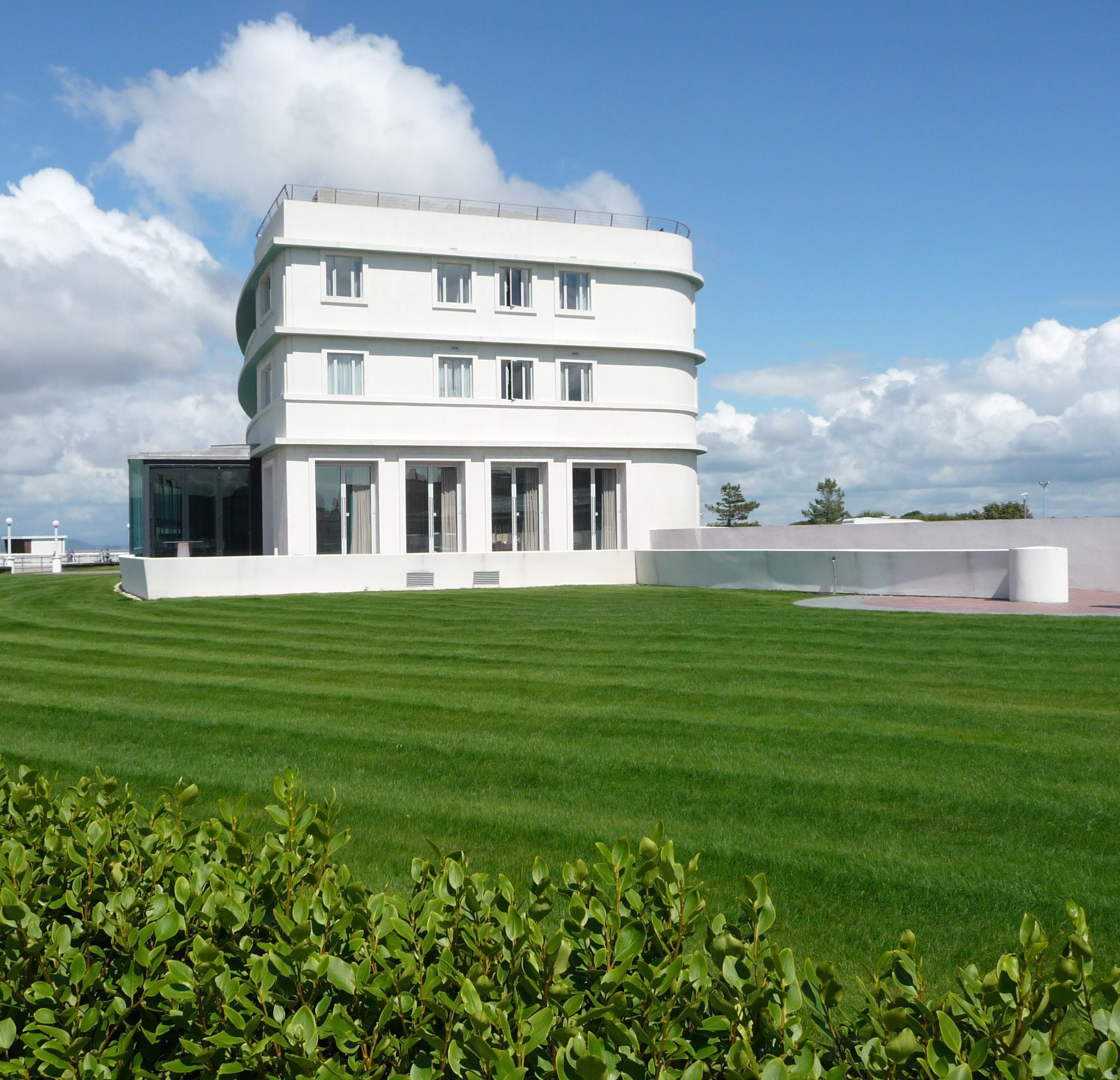 A modern white building with a flat roof and multiple windows, surrounded by a well-maintained green lawn and some bushes.