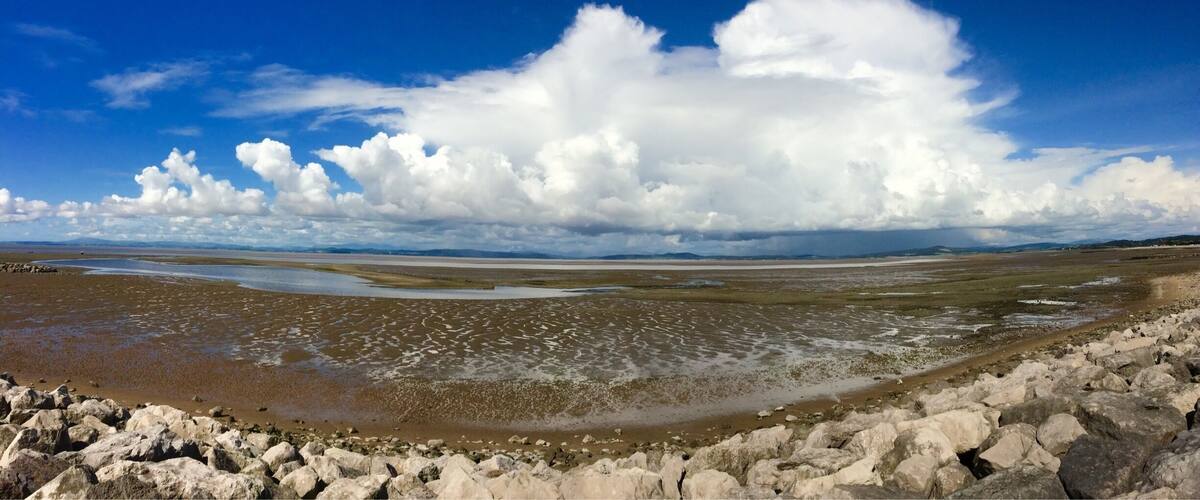 A coastal landscape with a rocky foreground, expansive mudflats, and a distant shoreline under a partly cloudy sky.