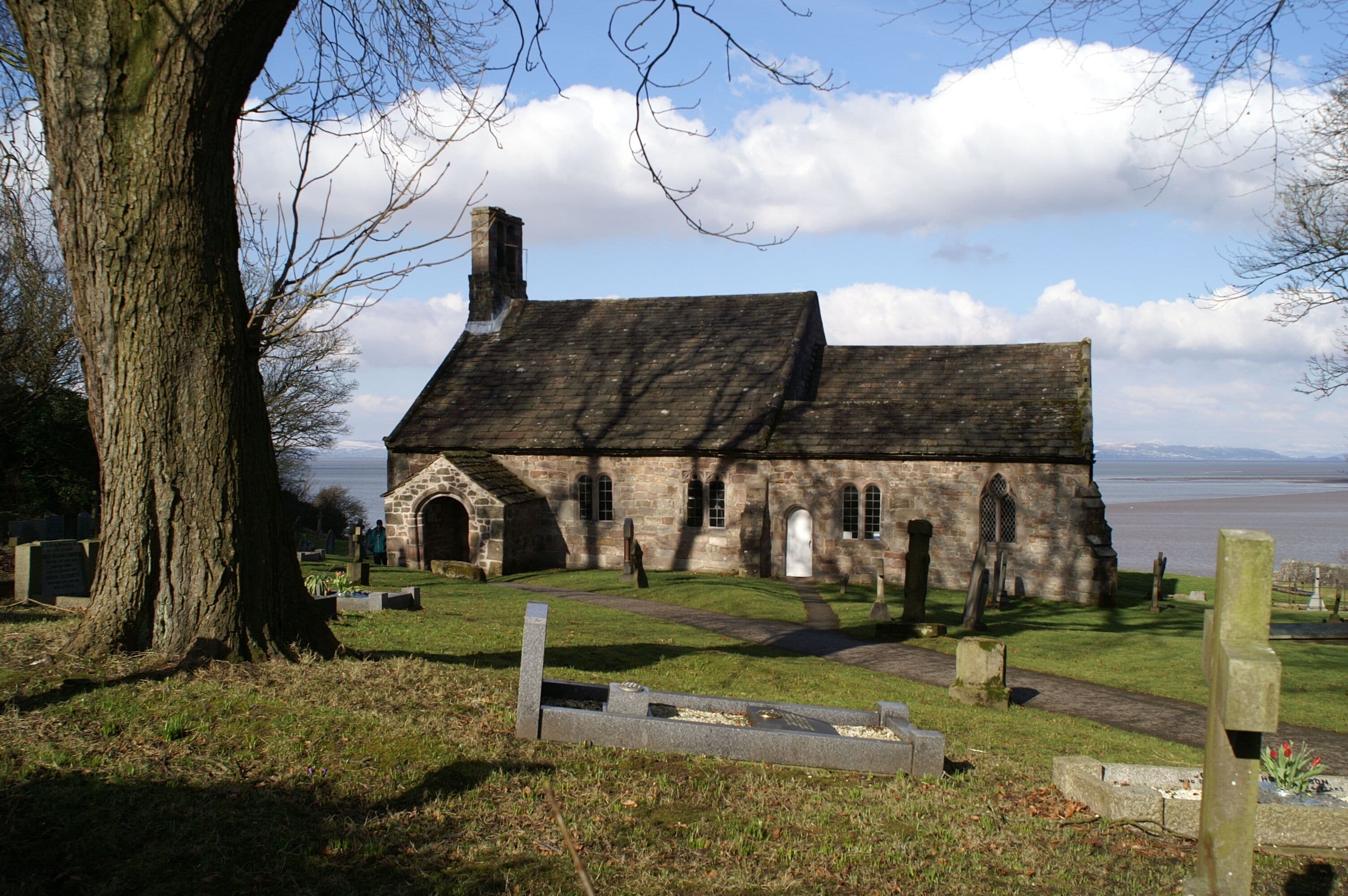 A stone church with a large tree in the foreground and a body of water in the background.