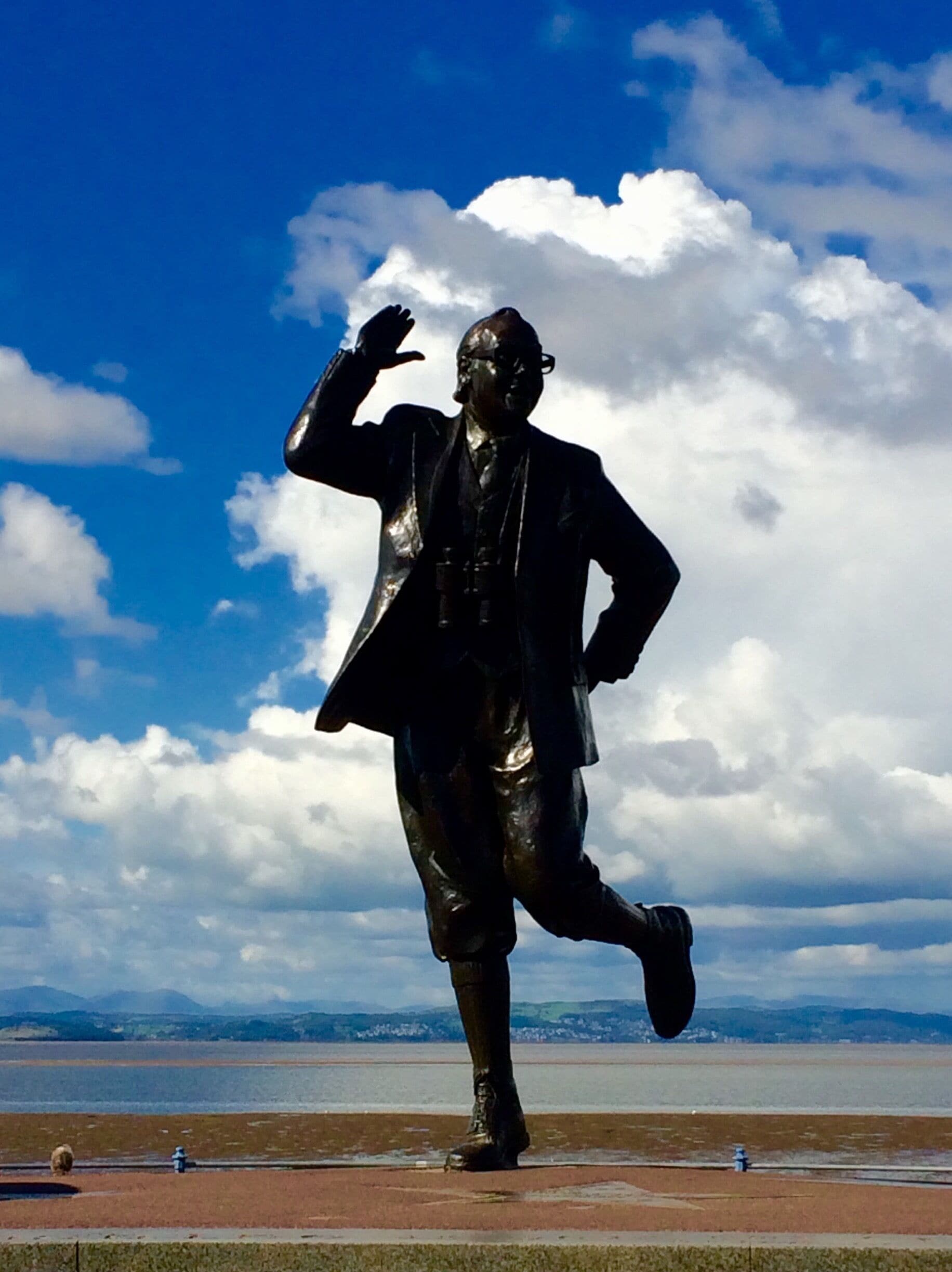 A statue of a man in a suit with one hand raised, set against a backdrop of a blue sky with clouds and a body of water in the distance.