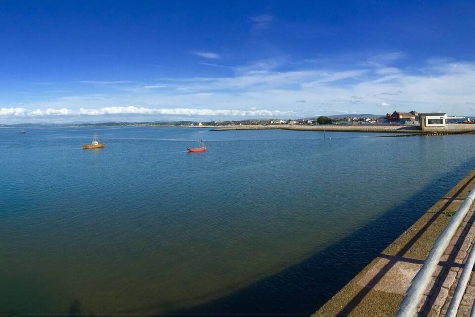 A calm waterfront with a paved walkway, boats, and distant buildings.