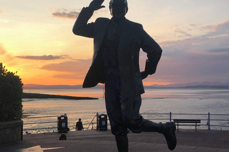 A statue of a man in a suit with a raised hand, standing on a star-shaped pavement with a sunset in the background.
