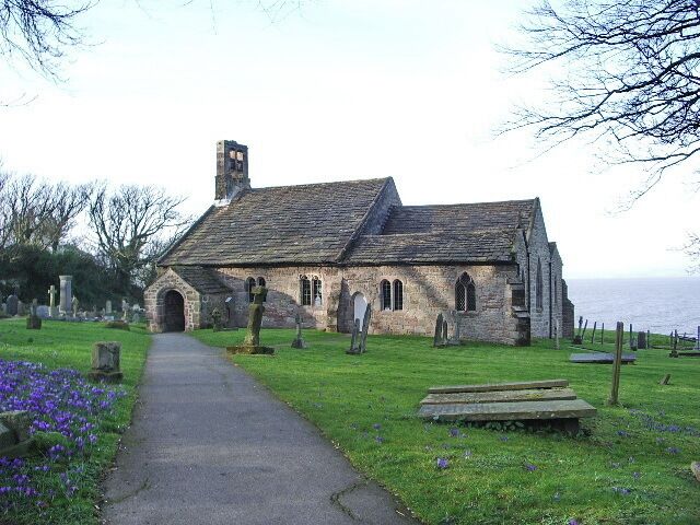 A stone church with a pathway leading to it, surrounded by a cemetery with headstones and purple flowers.