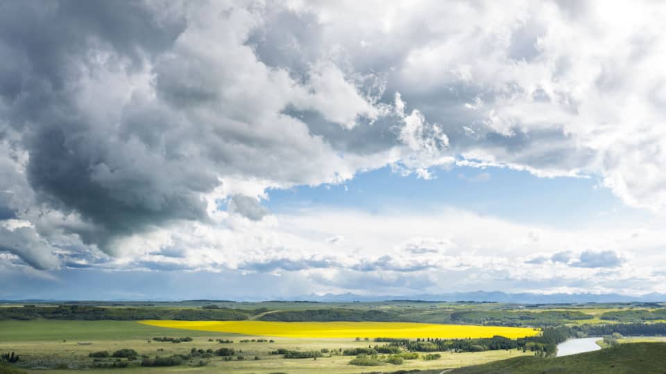 A landscape with a yellow field, green hills, a river, and a cloudy sky.