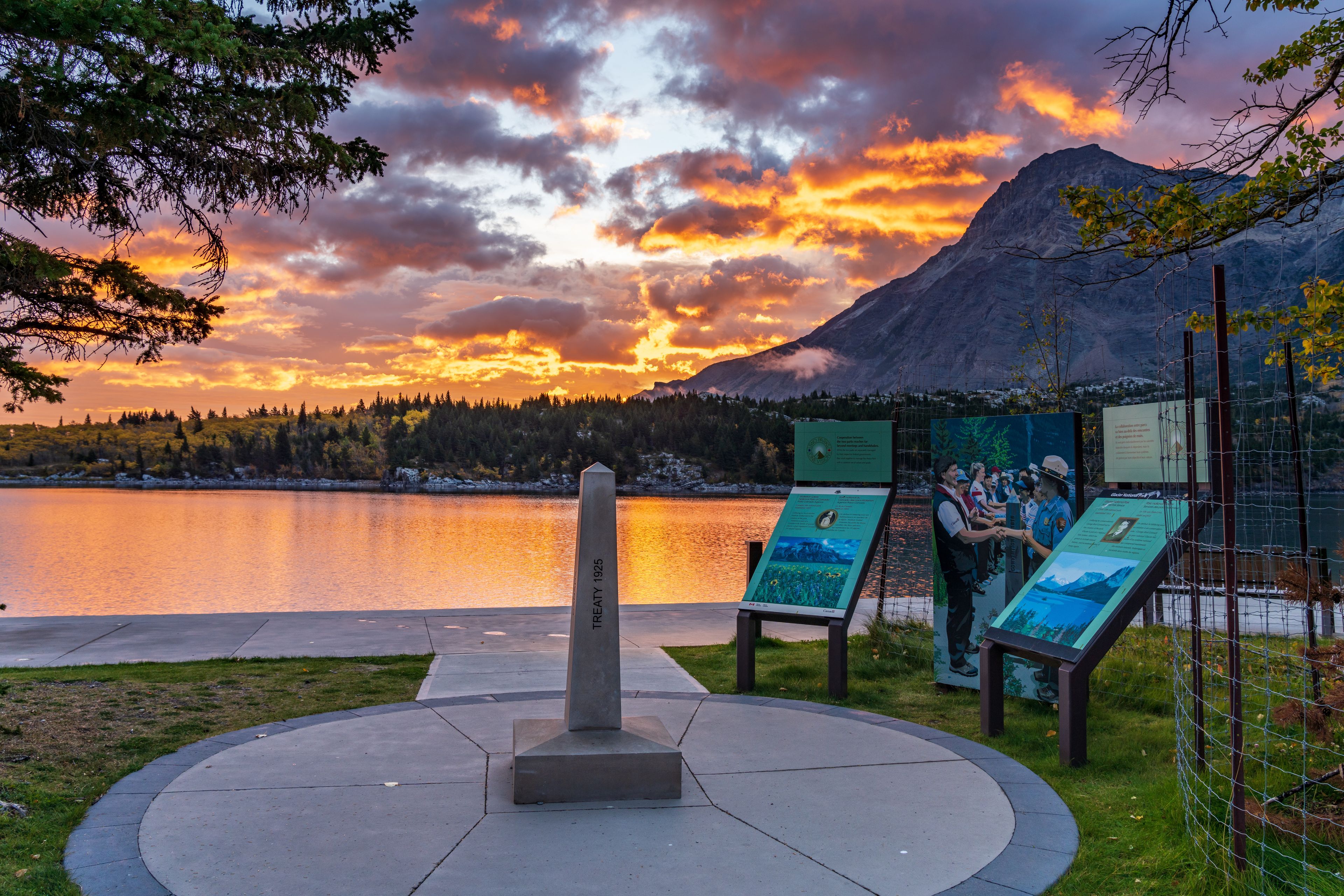 A monument with a plaque, a lake, a mountain, and informational signs.
