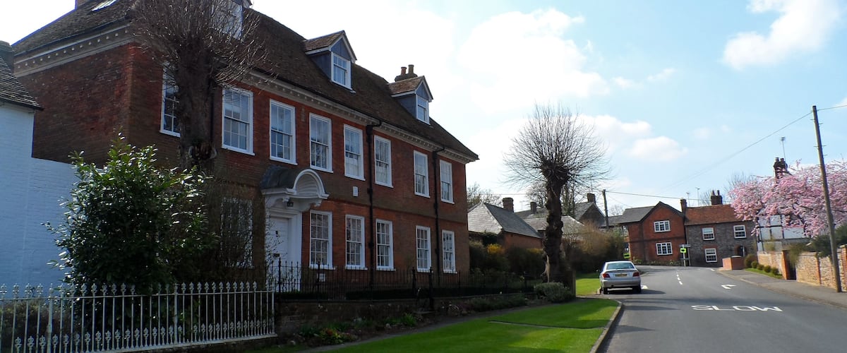 A quiet residential street with red brick houses, a well-maintained lawn, and a car driving on the road.