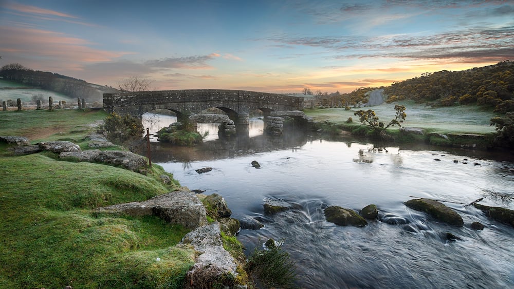 Eine Steinbrücke über einem Fluss mit einer grünen Landschaft und Bäumen im Hintergrund.