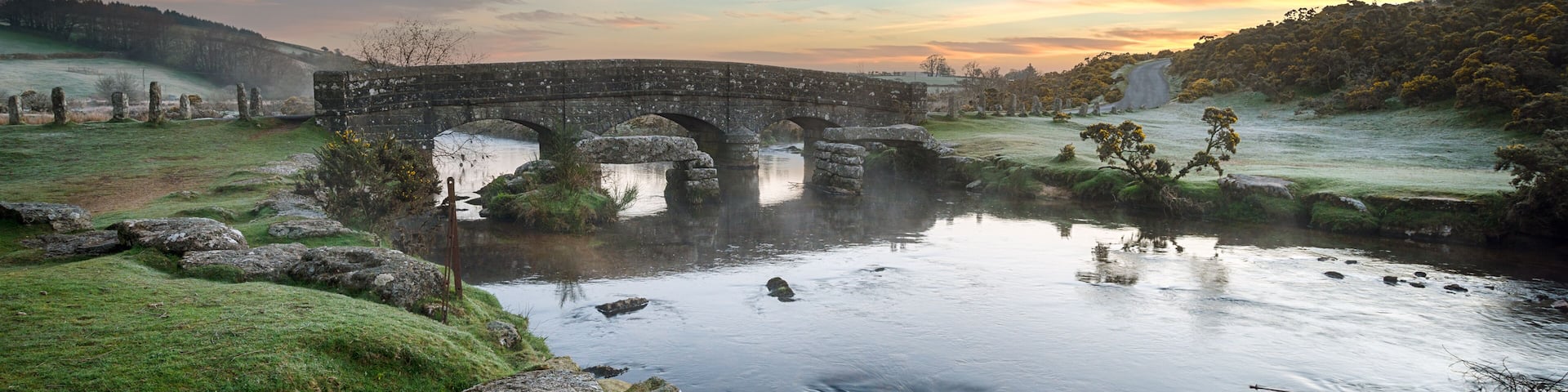 A stone bridge over a river with a grassy landscape and trees in the background.