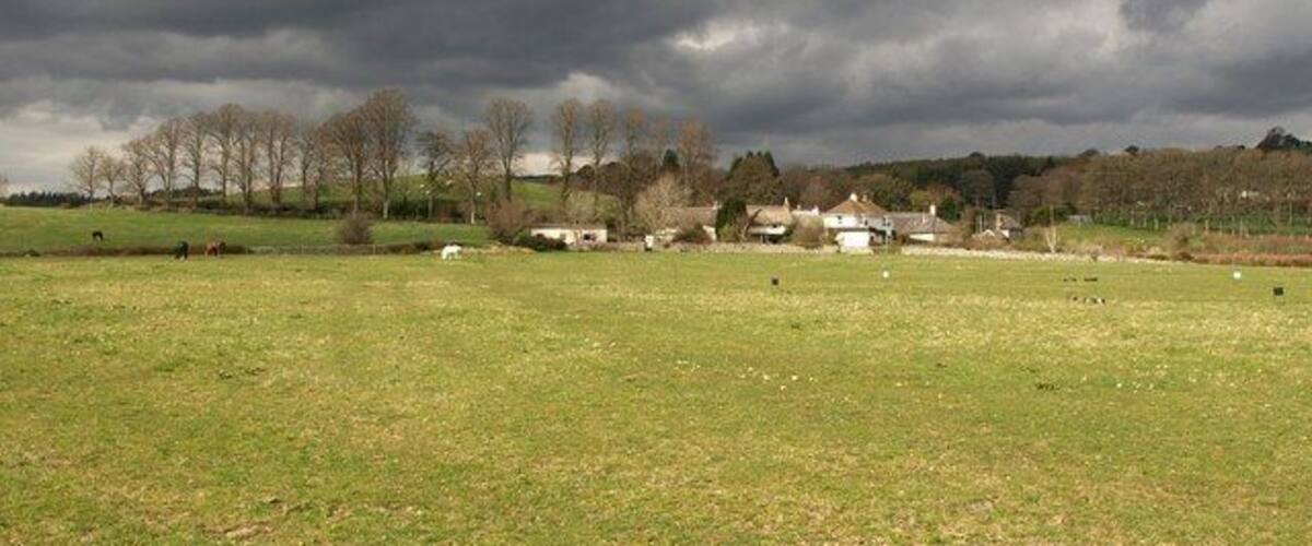 A rural landscape with a grassy field, a few scattered buildings, and a line of trees under a cloudy sky.