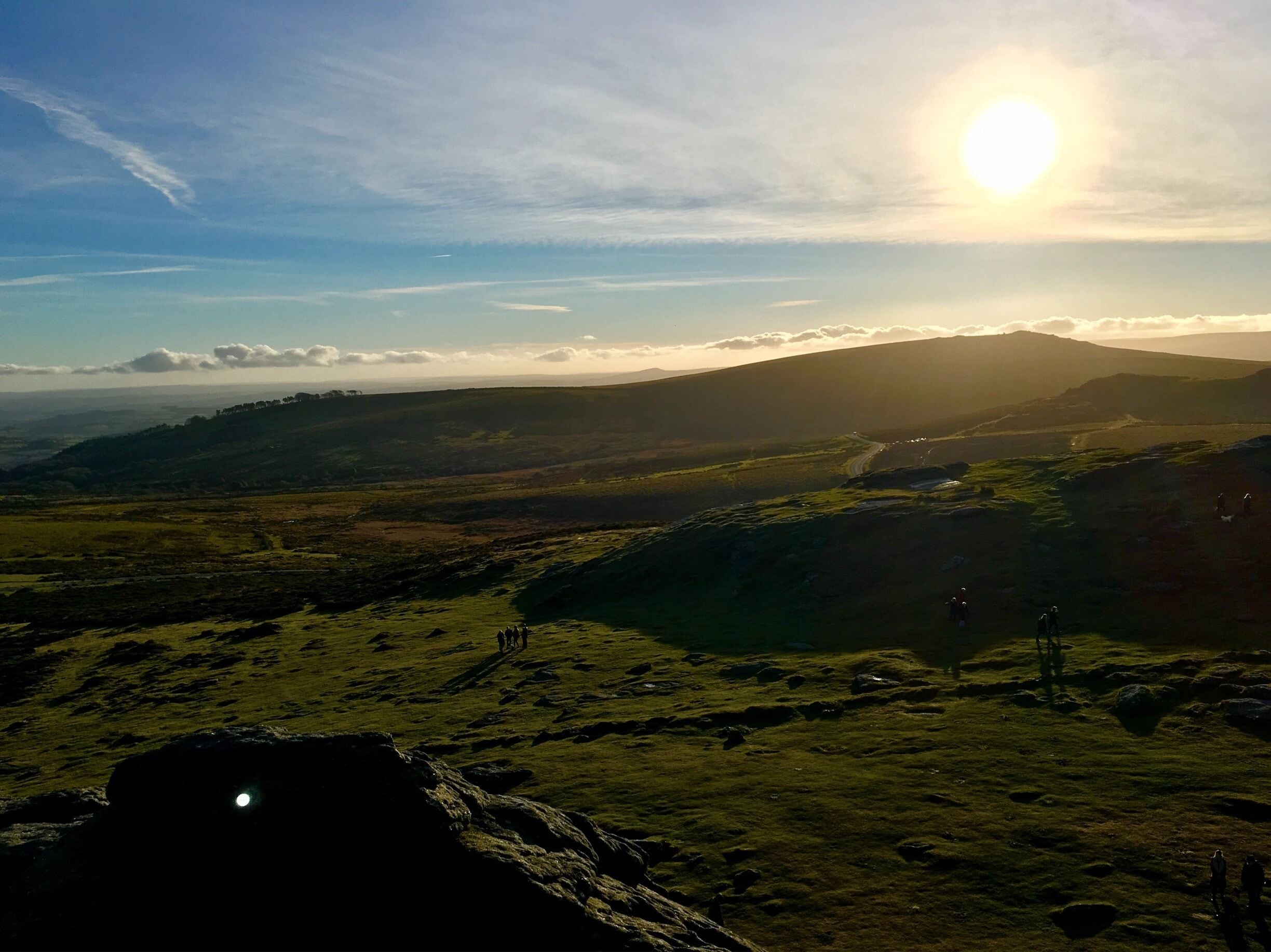 A mountainous landscape with a winding road, a small group of people, and a bright sun setting or rising in the background.