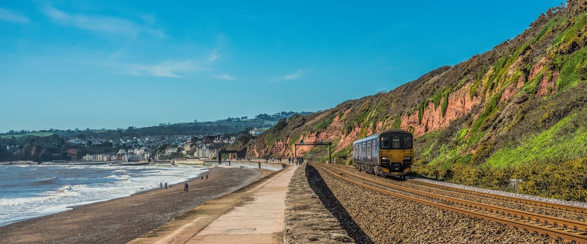 A coastal railway track with a train, beach, and houses in the background.