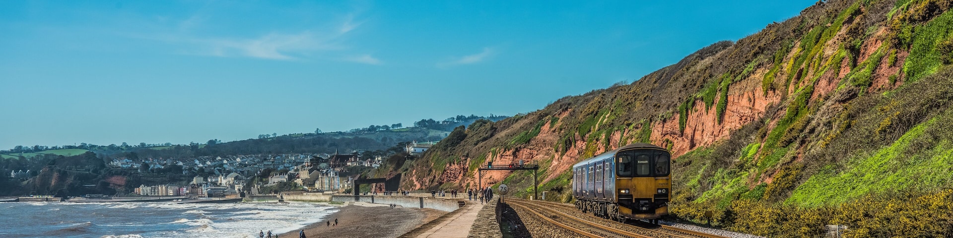 A coastal railway track with a train, beach, and houses in the background.