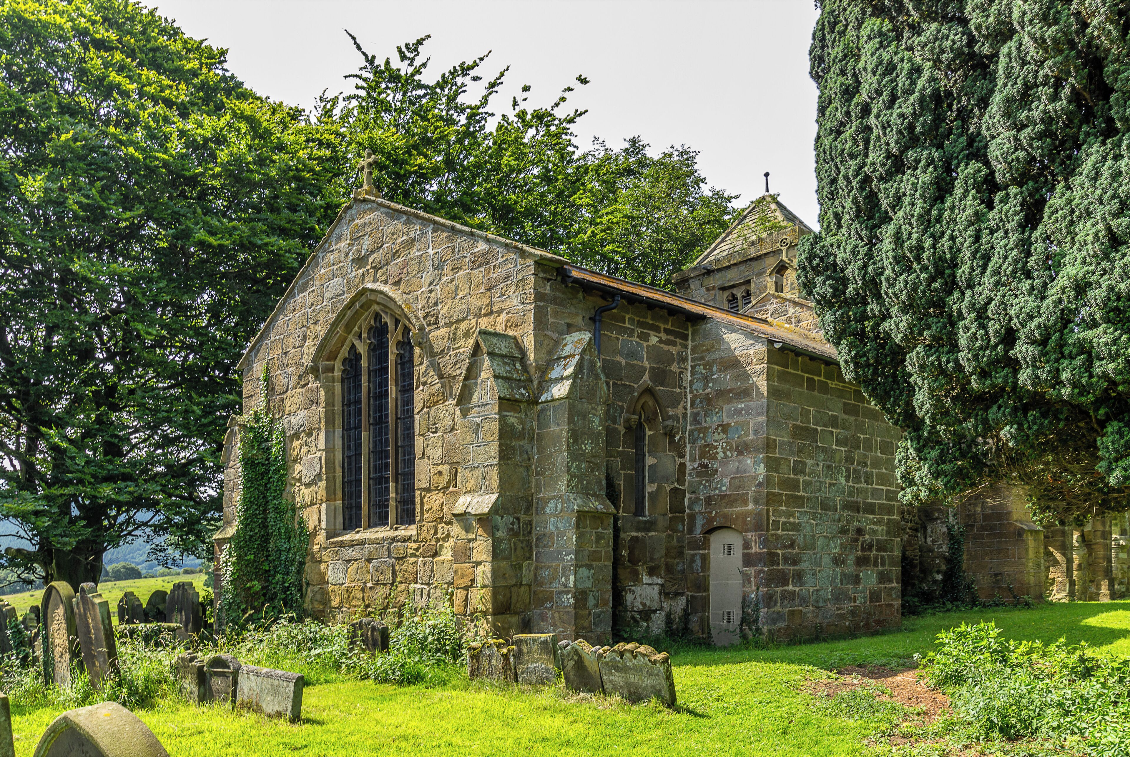 A small stone church with a pointed arch window and a bell tower, surrounded by lush greenery and a graveyard.