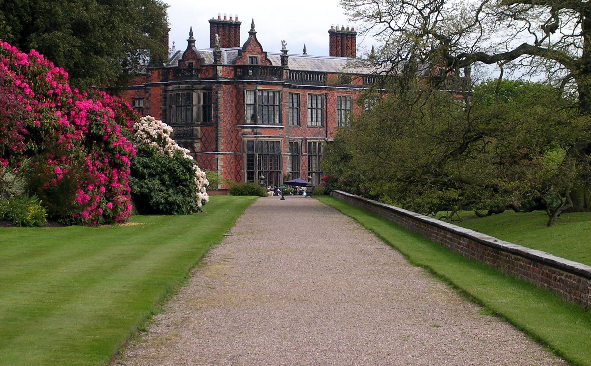 A historic building with a gravel pathway leading to it, surrounded by lush greenery and flowering bushes.