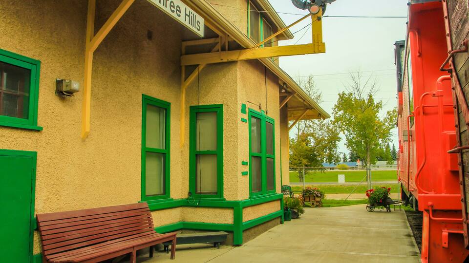 A train station with a sign reading 'C.N. Three Hills,' a bench, and a red train.
