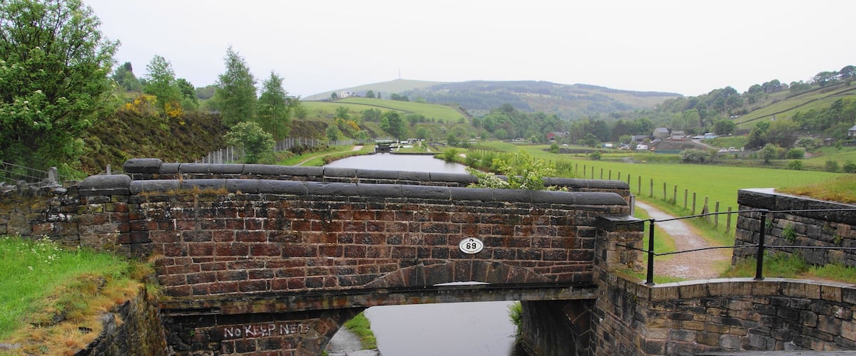 A stone bridge over a canal with a 'No Keep Under' sign, surrounded by greenery and hills.