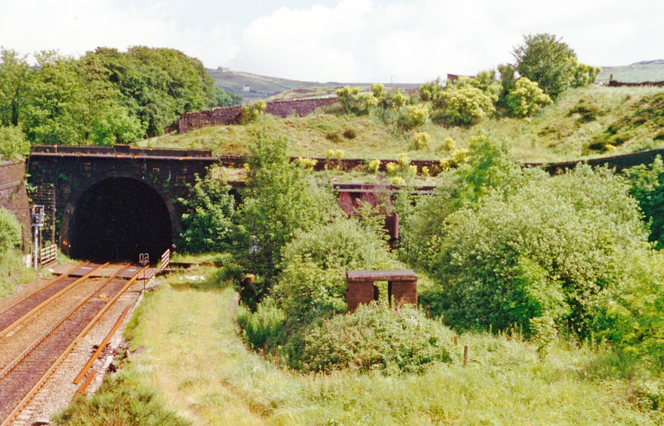 Western portal of Standedge Tunnel at Diggle. View NE from the A62 road, towards Huddersfield, Leeds etc.: ex-LNWR Manchester - Huddersfield - Leeds main line. Behind the camera is the site of the former Diggle station - see SE0008 : Site of former Diggle station, 1996. There are four tunnels here: the two tracks remaining constitute the main line, through the most recent (1894) tunnel bore, next to the right the original (1811) canal tunnel runs underneath here. Further over to the right are the original (1848 and 1871) single-line rail tunnels, still extant but with no track although one is available for emergency road vehicles.