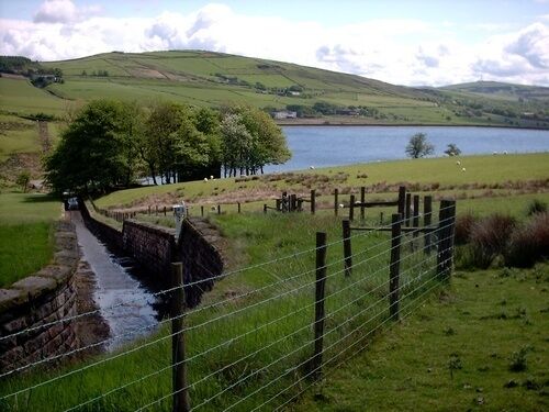 A narrow water channel, a fenced area, and a lake in a hilly landscape.