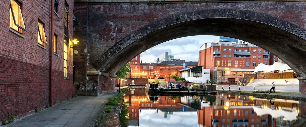 A canal under a stone bridge with brick buildings and a museum in the background.