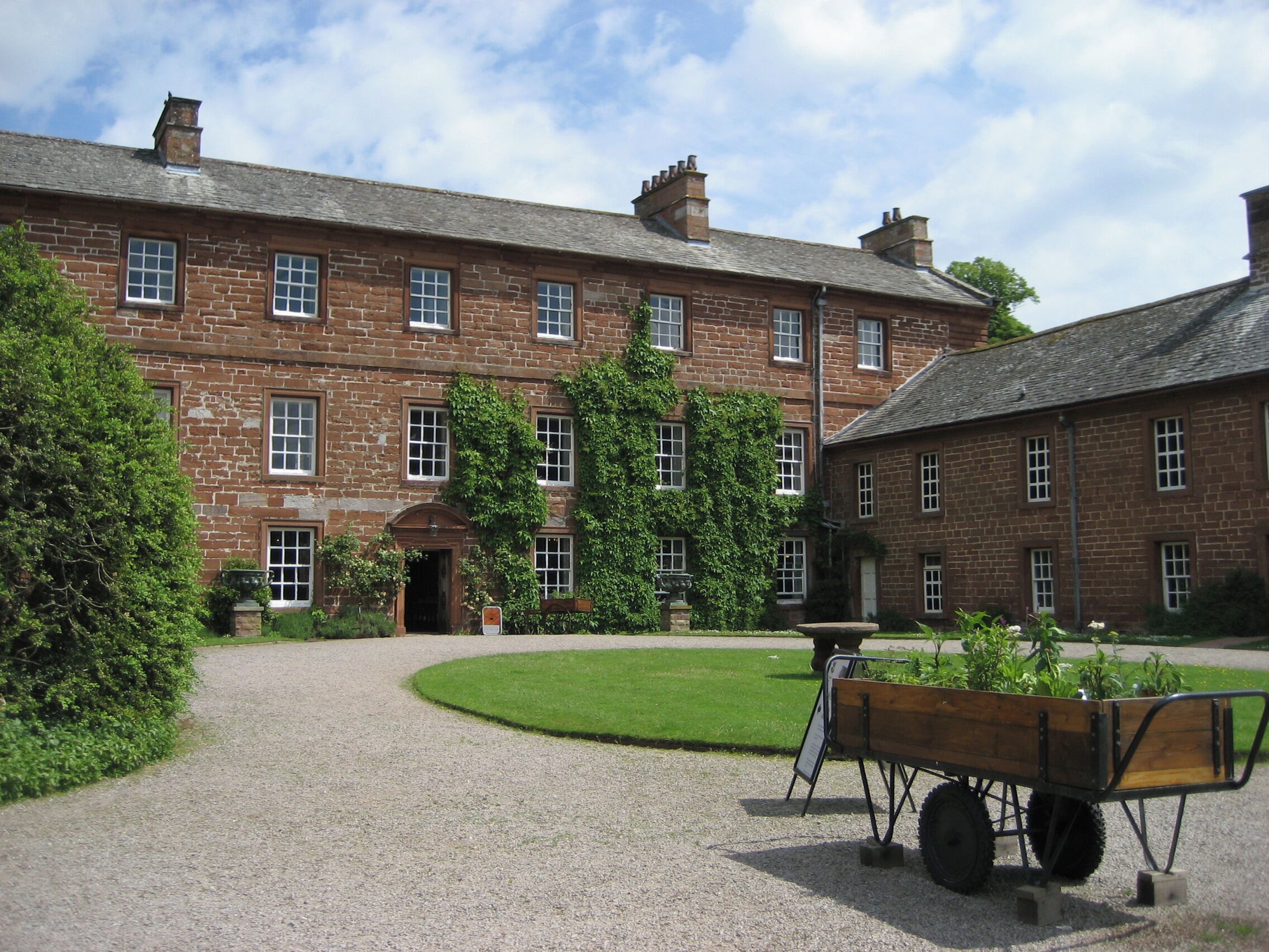A historic brick building with a gravel courtyard, a wooden wheelbarrow, and lush greenery.