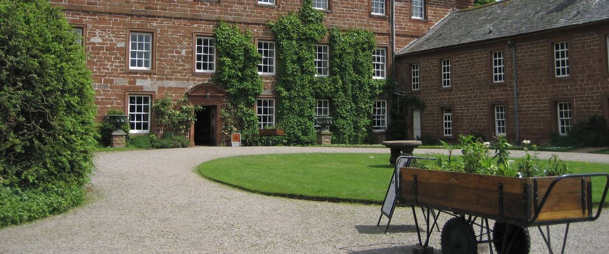 A historic brick building with a gravel courtyard, a wooden wheelbarrow, and lush greenery.