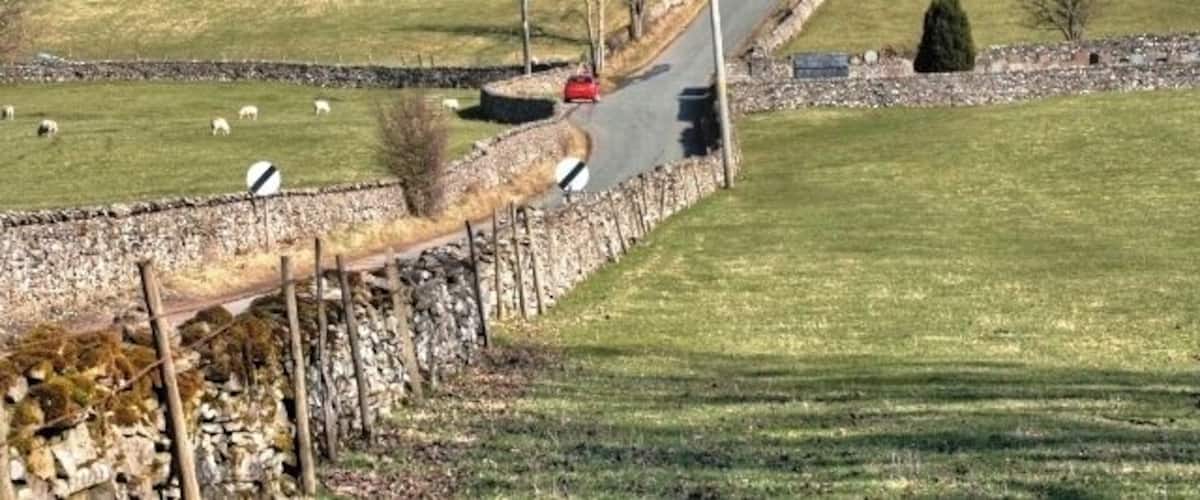 A rural road curving through a stone wall, with sheep grazing in the fields and a red car on the road.