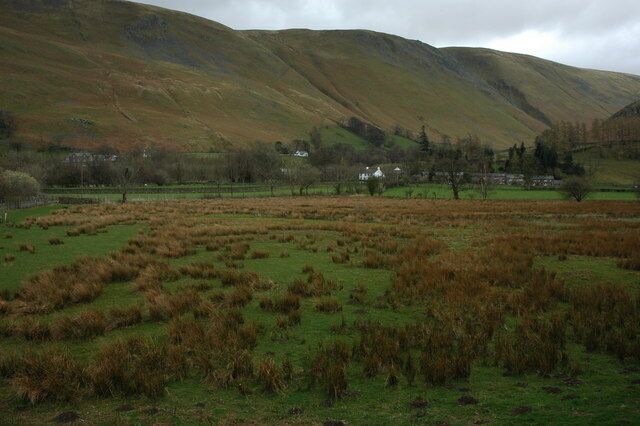 Paisagem rural com colinas ondulantes, um campo com vegetação castanha e um pequeno conjunto de casas ao longe.