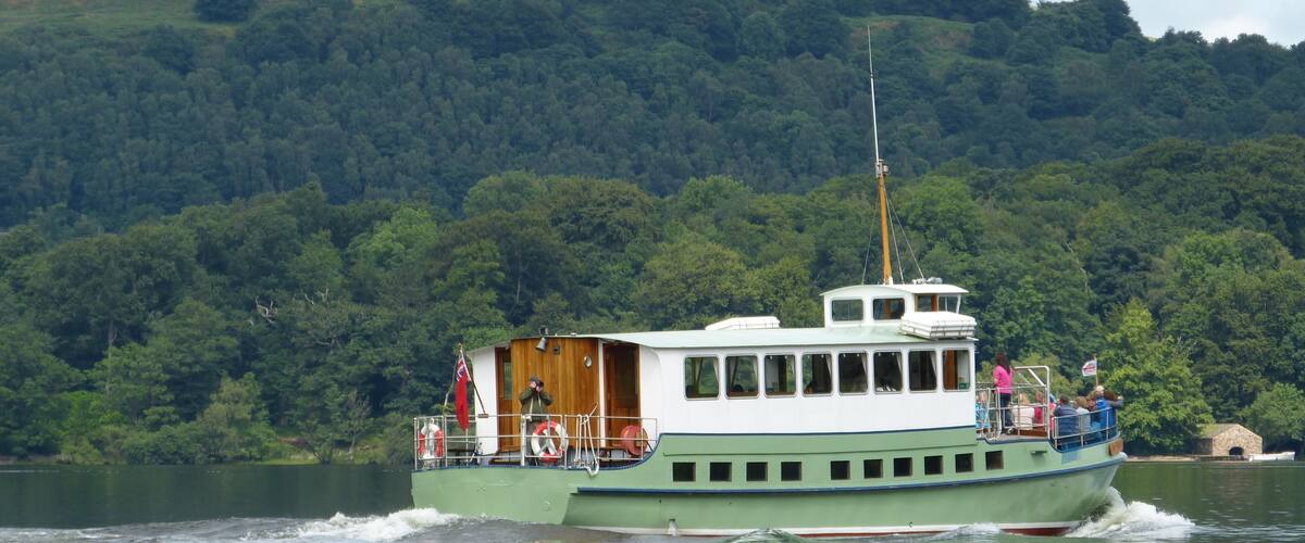 A green and white boat with passengers on a lake surrounded by dense forest and hills.