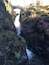 Une cascade étroite qui serpente à travers un défilé rocheux, avec des rochers couverts de mousse et un pont de pierre en arrière-plan.