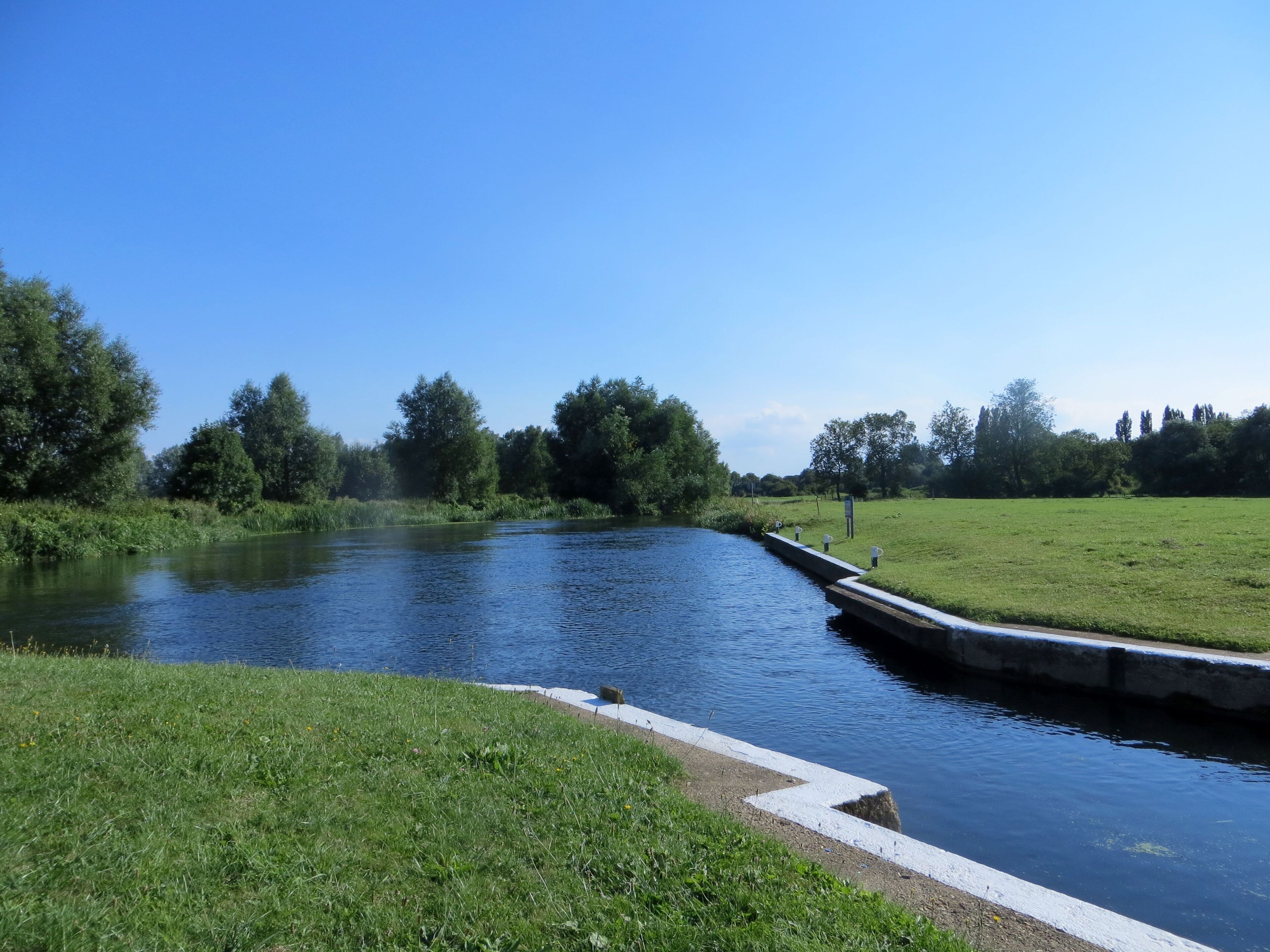 A calm river with a concrete embankment, surrounded by lush green fields and trees under a clear blue sky.
