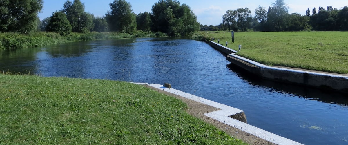 A calm river with a concrete embankment, surrounded by lush green fields and trees under a clear blue sky.