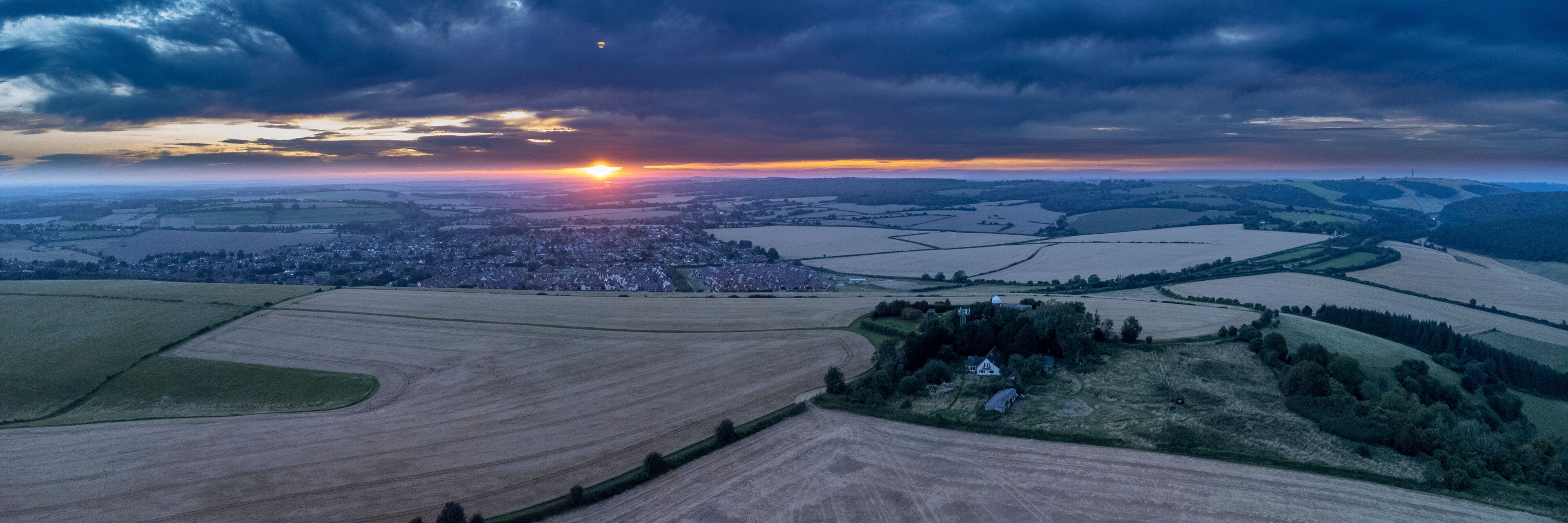 A rural landscape with fields, a village, and a cloudy sky at sunset.