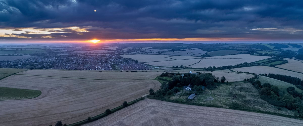 A rural landscape with fields, a village, and a cloudy sky at sunset.