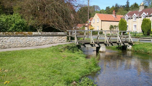 A wooden bridge over a calm river, surrounded by greenery and stone walls, with houses in the background.