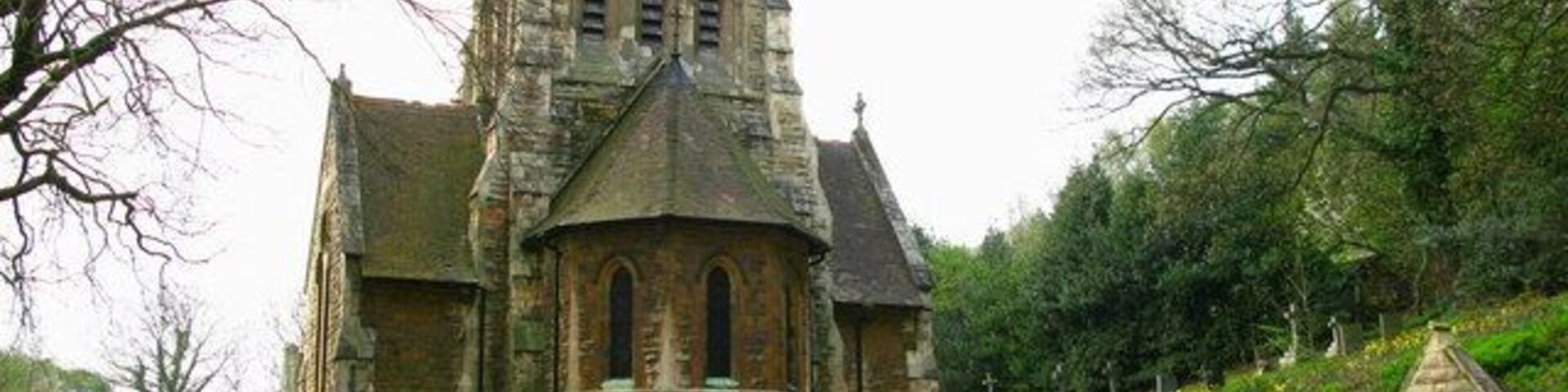 Uma igreja de pedra histórica com uma torre imponente e janelas em arco, rodeada por vegetação exuberante e um caminho.