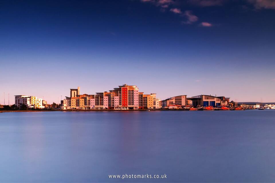 A waterfront cityscape with a prominent red and white building, a calm river, and a clear sky.