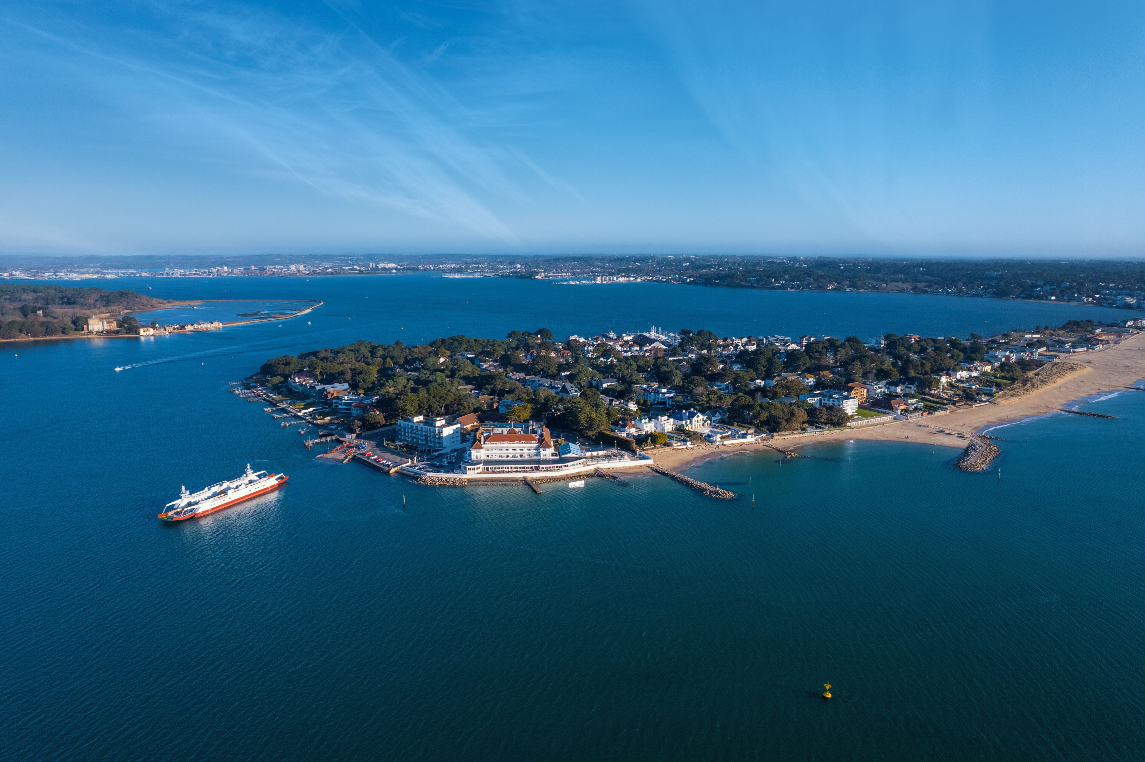 An aerial view of a coastal town with a prominent red ship docked at the pier, surrounded by dense greenery and a clear blue sky.
