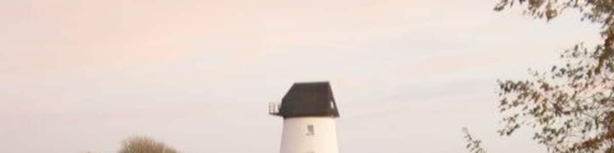 Ein weißer Leuchtturm mit schwarzer Spitze, ein kleines weißes Gebäude und ein Teich, der die Struktur widerspiegelt.