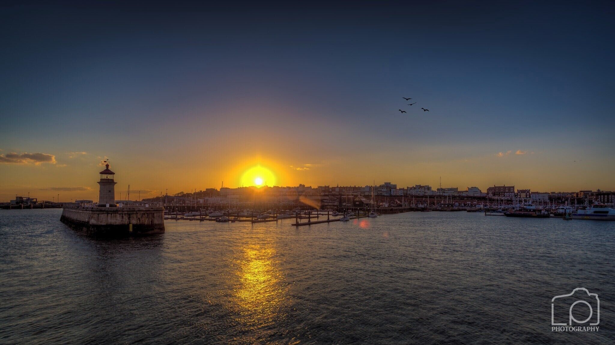 A sunset over a harbor with a lighthouse, boats, and a city skyline.
