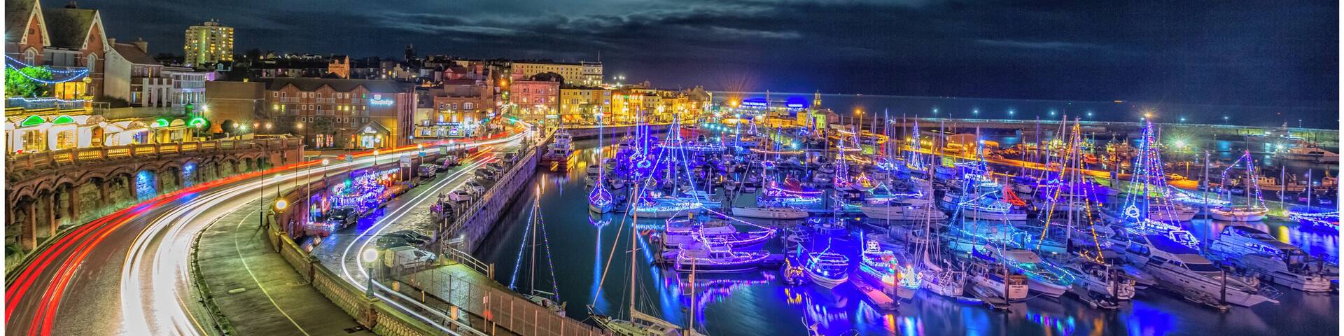 A marina at night with boats illuminated by colorful lights, a curved road with light trails, and a cityscape in the background.