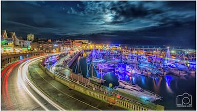 Uma marina à noite, com barcos iluminados por luzes coloridas, uma estrada curva com rastros de luz e um horizonte urbano ao fundo.