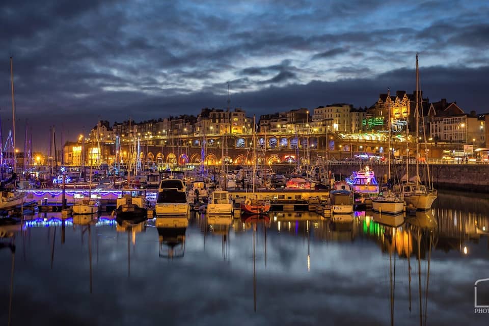 Una marina de noche con barcos amarrados y edificios iluminados al fondo.