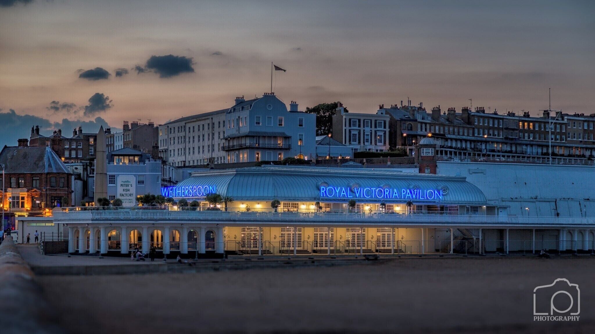 The Royal Victoria Pavilion with a blue neon sign, Peter's Fish Factory, and surrounding historic buildings.
