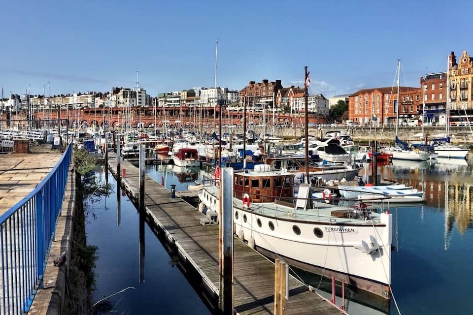Una marina con varios barcos amarrados a lo largo de un muelle de madera, rodeada de edificios y un cielo azul despejado.