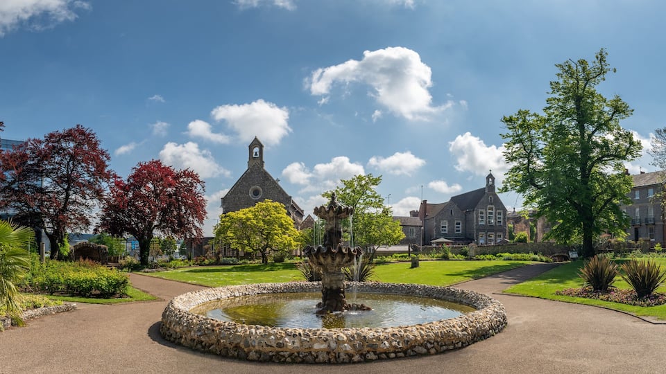 A park with a fountain, a church, and surrounding buildings.