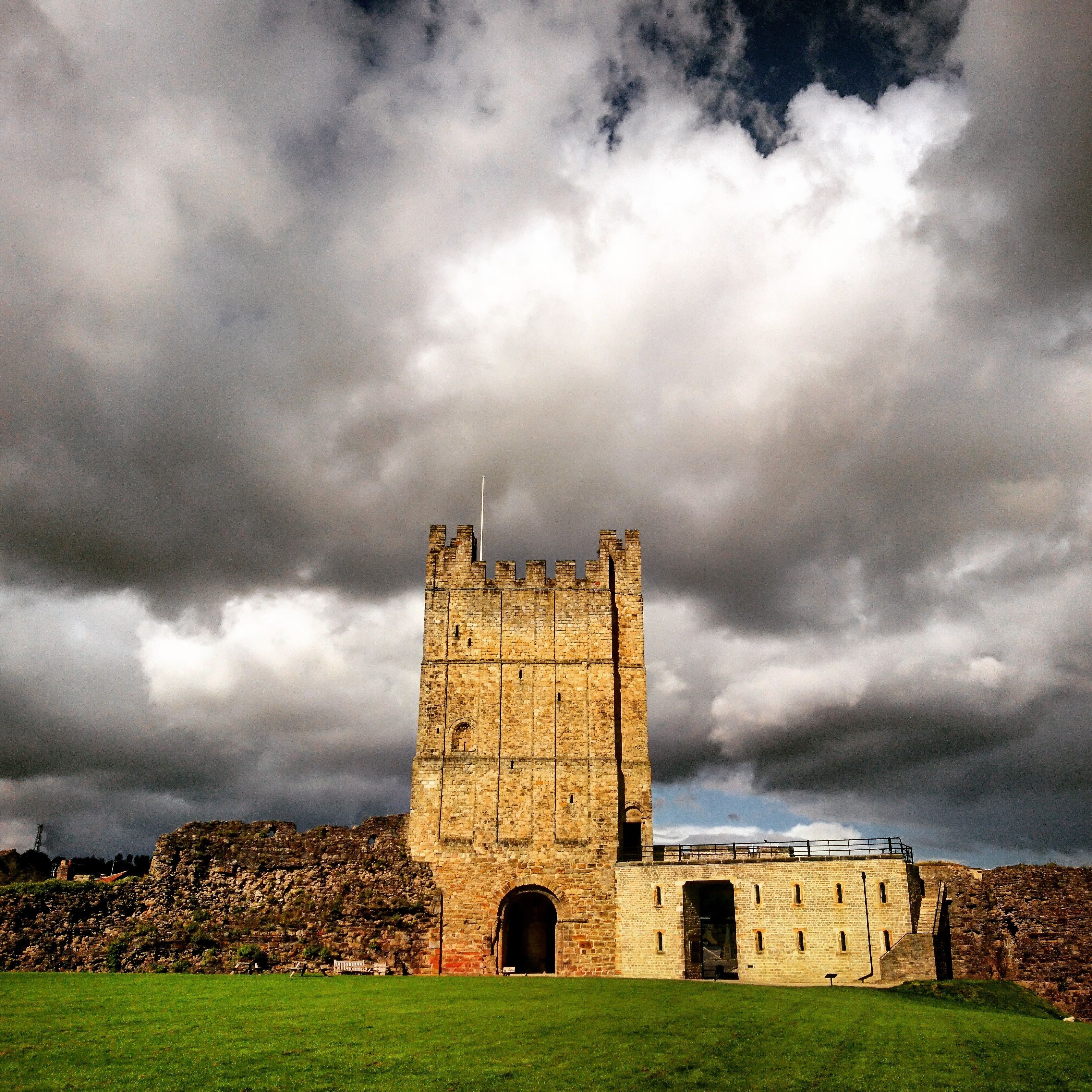 A historic stone tower with a large arched entrance, surrounded by a grassy field and a rocky hill in the background.